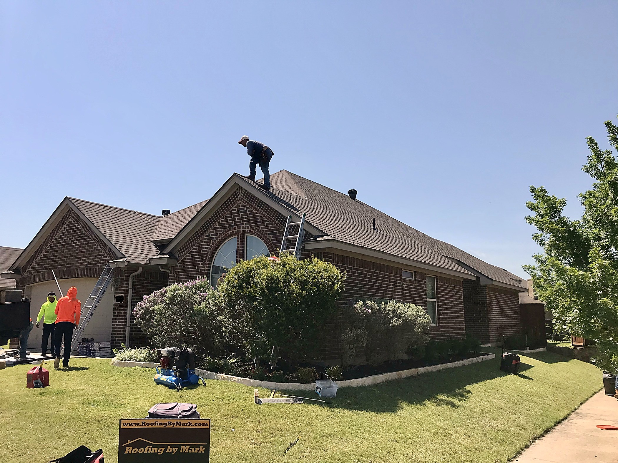 Crew and ladders on the lawn of a brick home, with a Roofing by Mark yard sign in front