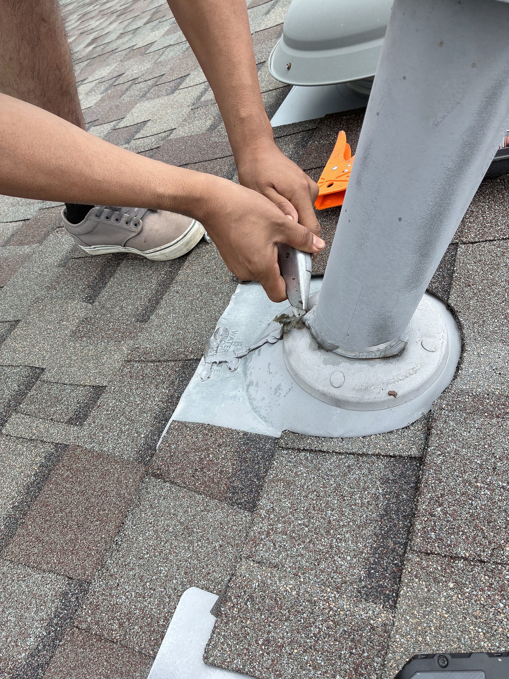 Hands sealing the base of a vent pipe on a shingle roof