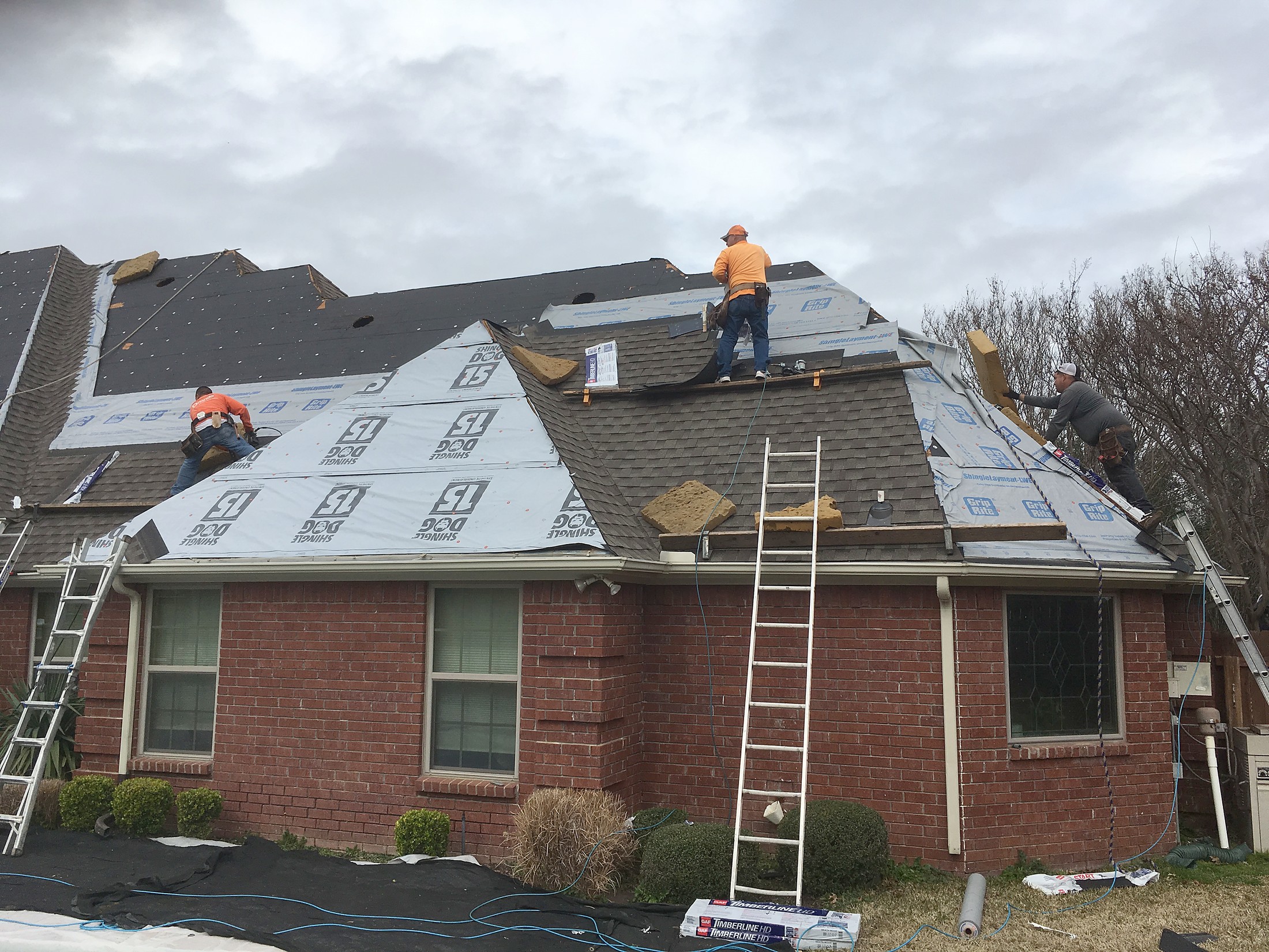 A crew on ladders replacing the roof of a red brick home, with new underlayment and shingles going down